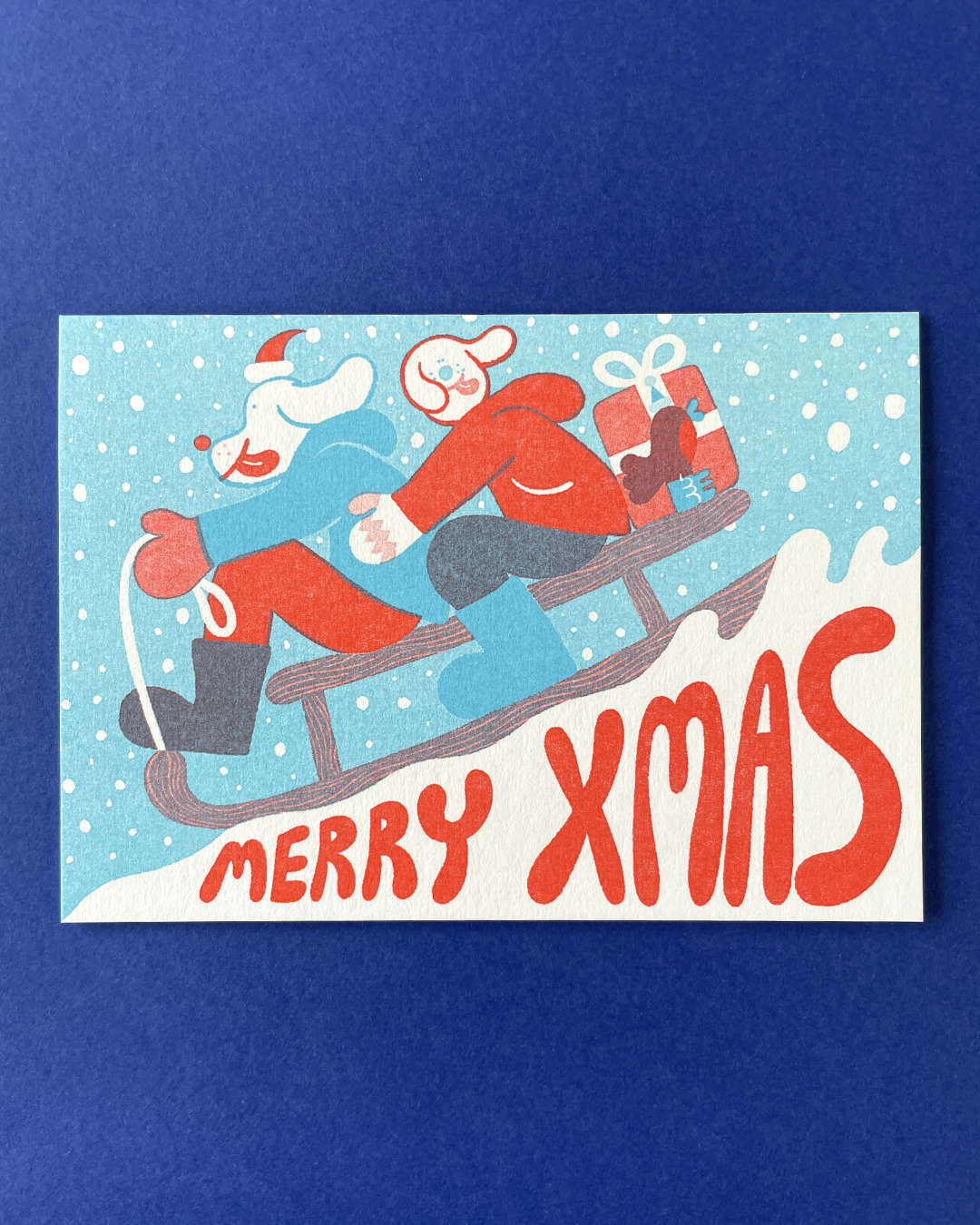 YUK FUN Christmas card with two dog people sledding down a snowy hill, text 'Merry Xmas' on a blue background.