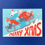 YUK FUN Christmas card with two dog people sledding down a snowy hill, text 'Merry Xmas' on a blue background.