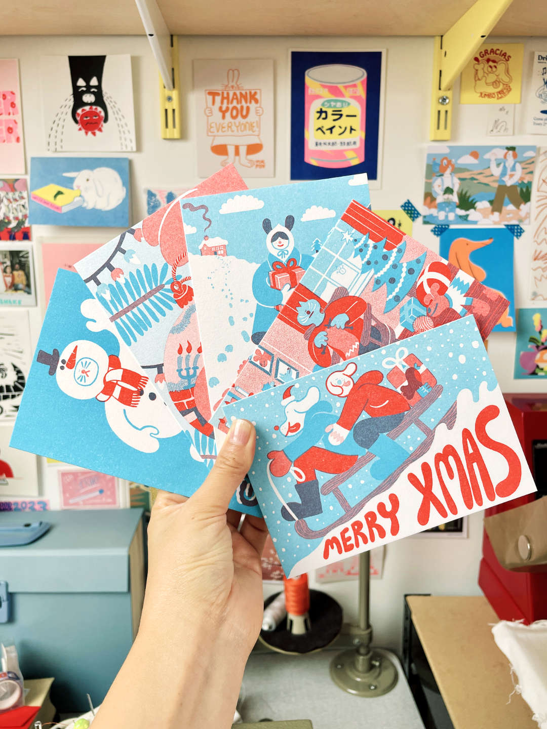 Hand holding YUK FUN Christmas-themed cards with colorful designs against a wall of posters.