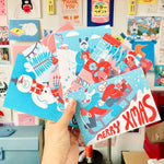 Hand holding YUK FUN Christmas-themed cards with colorful designs against a wall of posters.